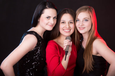 Portrait of three elegant beautiful girls posing with microphone happy smiling in colorful light of spotlights